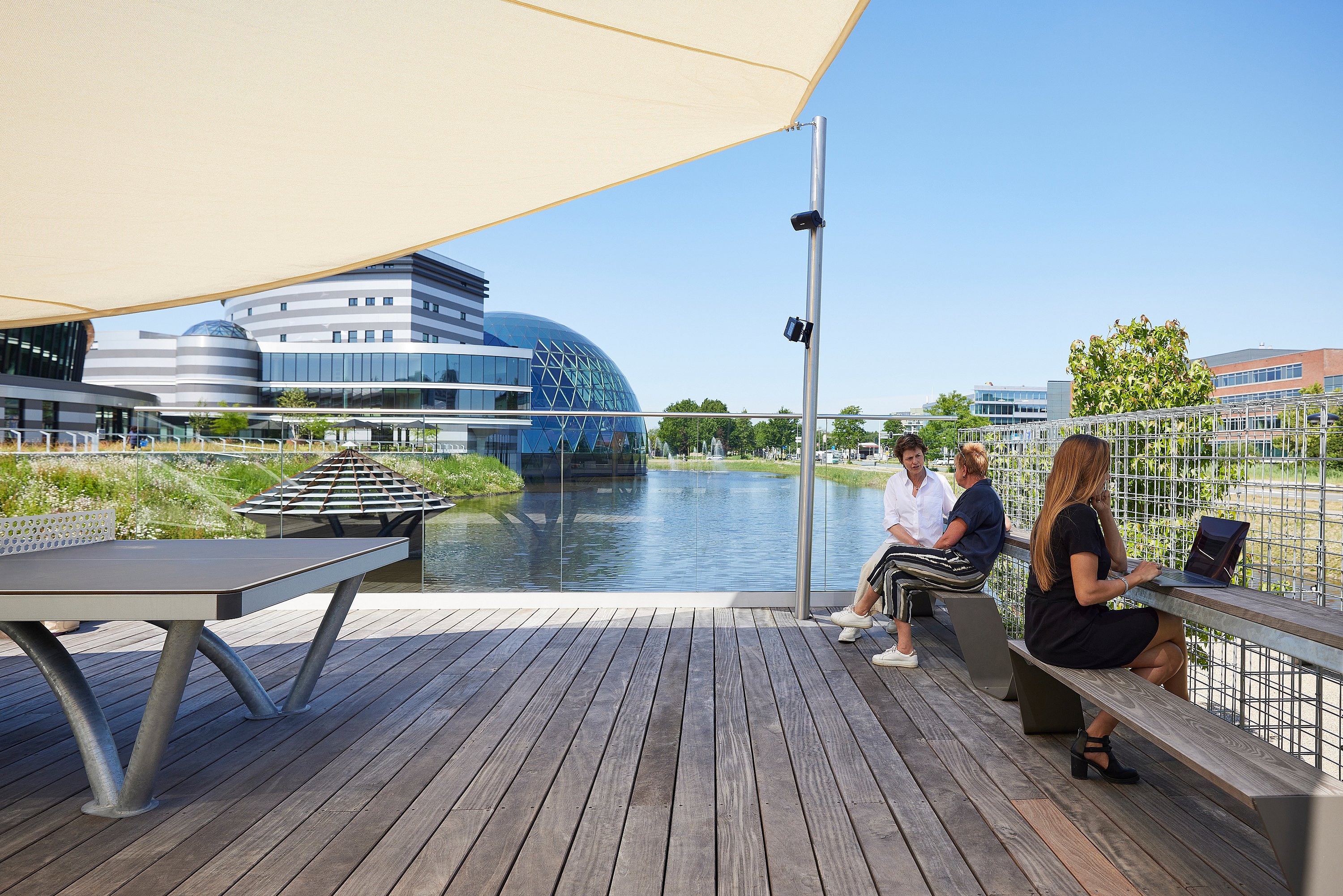 Sur une terrasse au bord de l’eau, une femme travaille à une longue table en bois, tandis que deux autres discutent sous une grande voile d’ombrage, avec les bâtiments du campus AFAS en arrière-plan.