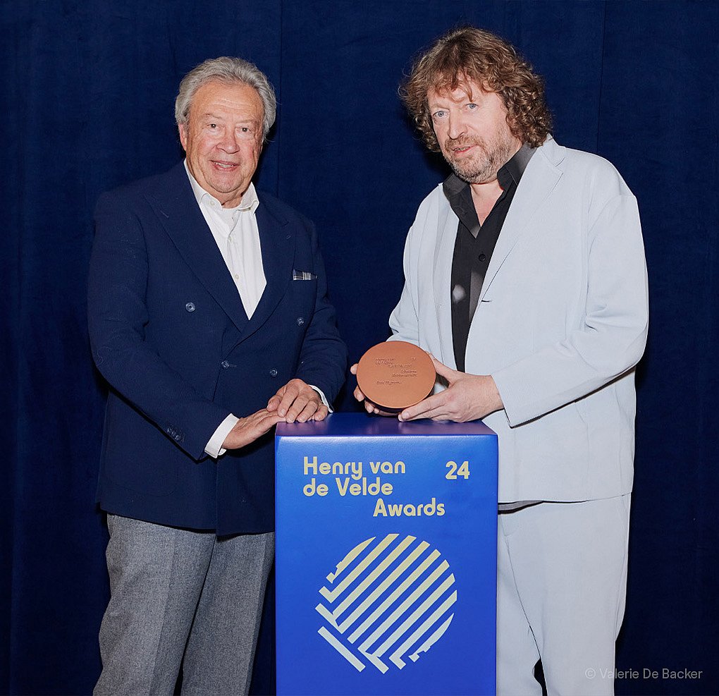 Portret van Dirk Wynants die buiten glimlachen naast een groot rood sculpturaal paneel, wat zijn Henry van de Velde Lifetime Achievement Award-erkenning markeert.