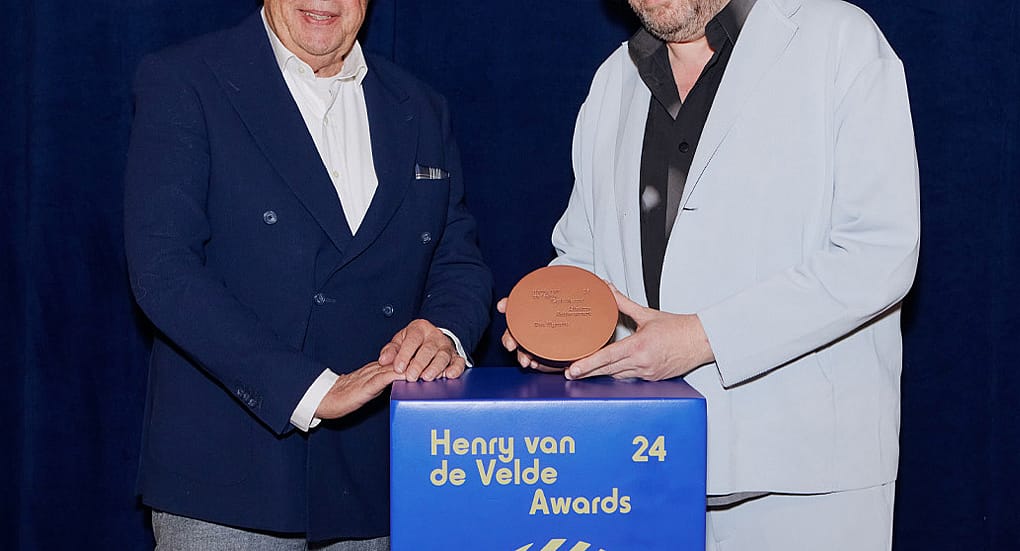 Portret van Dirk Wynants die buiten glimlachen naast een groot rood sculpturaal paneel, wat zijn Henry van de Velde Lifetime Achievement Award-erkenning markeert.