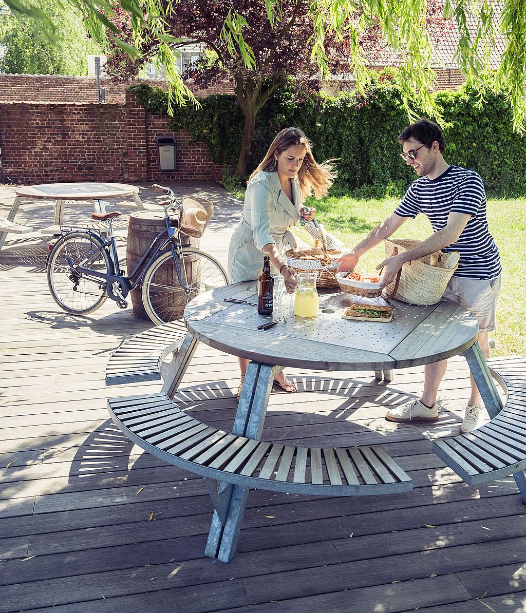 Two people unpacking food from picnic baskets onto a round outdoor Gargantua table on a wooden deck, with a bicycle parked beside them in a sunny garden.