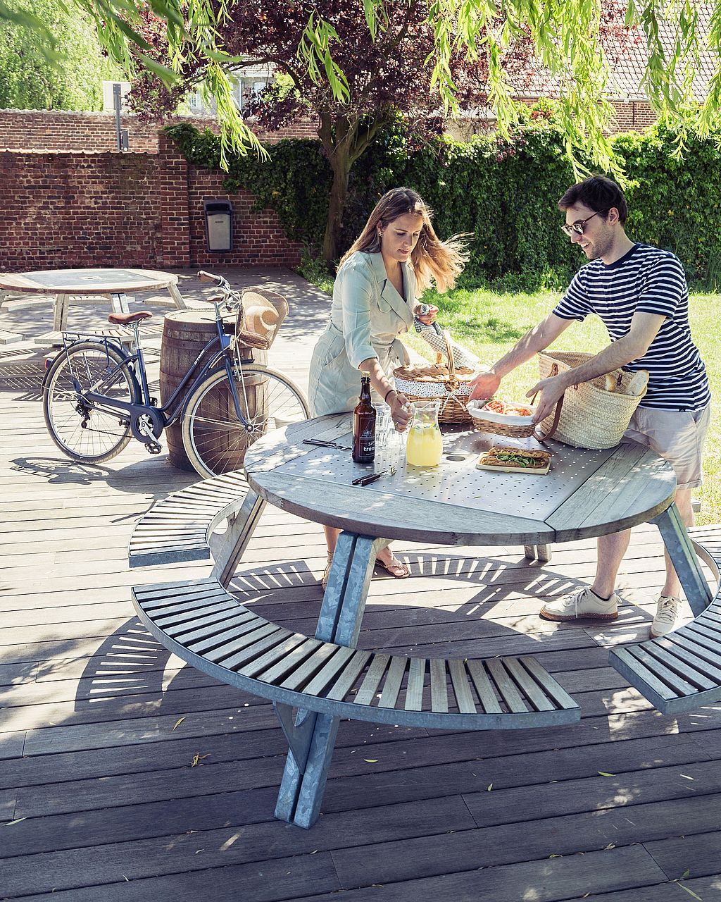 Twee mensen komen met de fiets aan bij een ronde picknicktafel voor buiten op een houten terras, met één persoon met een mand onder overhangende boomtakken.
