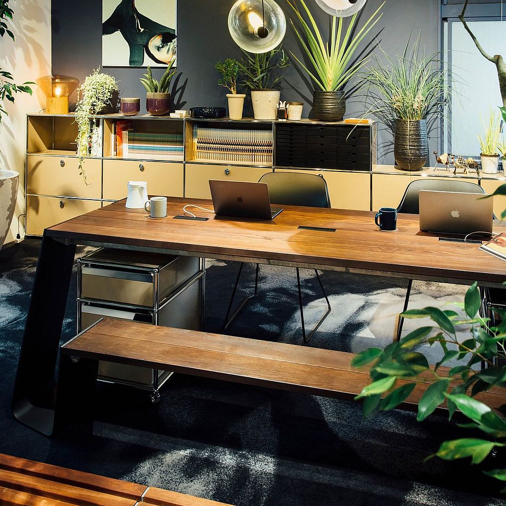 Warm, plant-filled office interior with a long wooden meeting table, bench seating, open laptops, mugs, and storage units, creating a relaxed and biophilic workspace atmosphere.