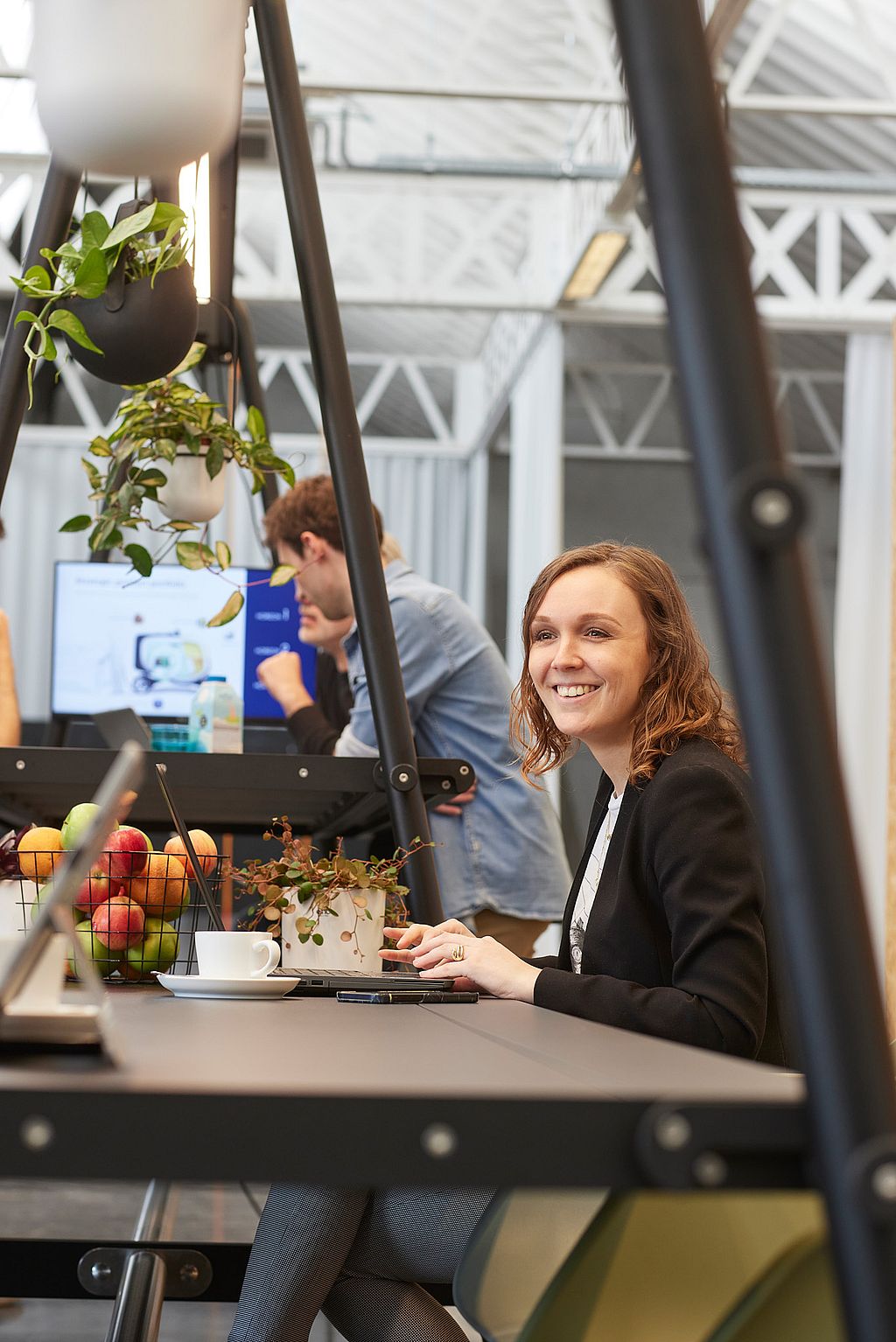 Woman smiling while seated at a shared indoor workstation framed by hanging plants and black structural supports, with coworkers blurred in the background.