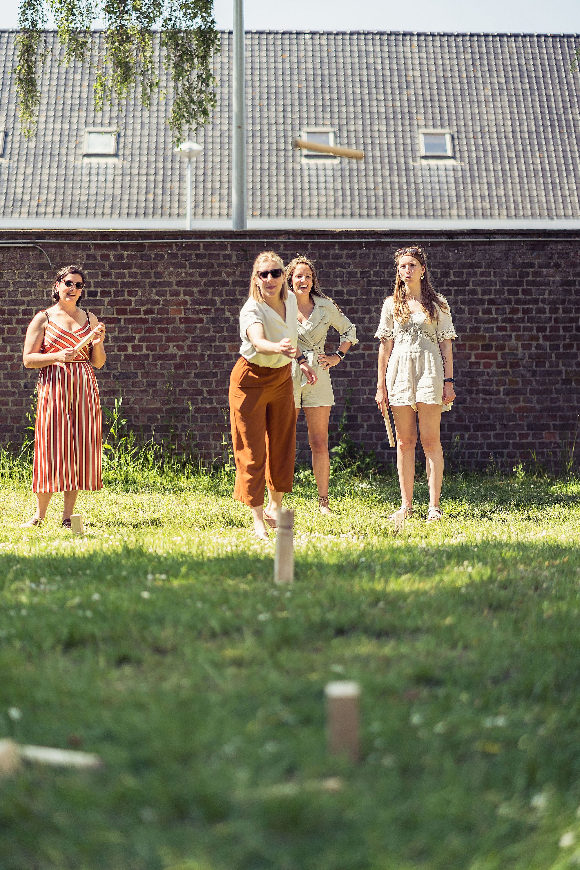 Four women playing Kubb on a grassy lawn, with one wooden baton flying toward the wooden blocks.