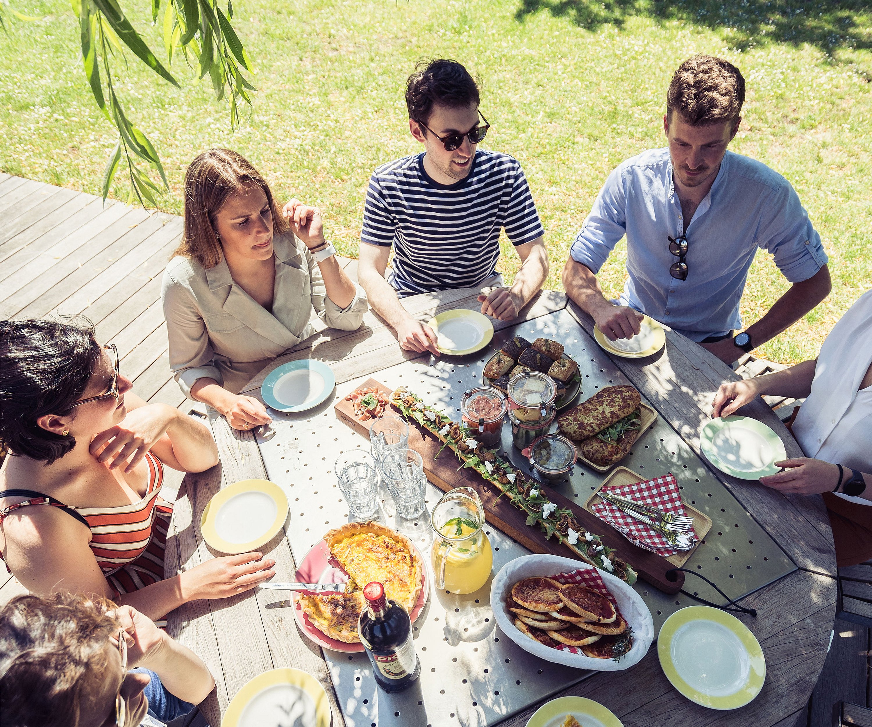 Overhead view of six people seated around a round picnic table covered with quiche, sandwiches, drinks and shared picnic dishes.
