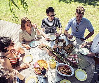 Overhead view of six people seated around a round picnic table covered with quiche, sandwiches, drinks and shared picnic dishes.