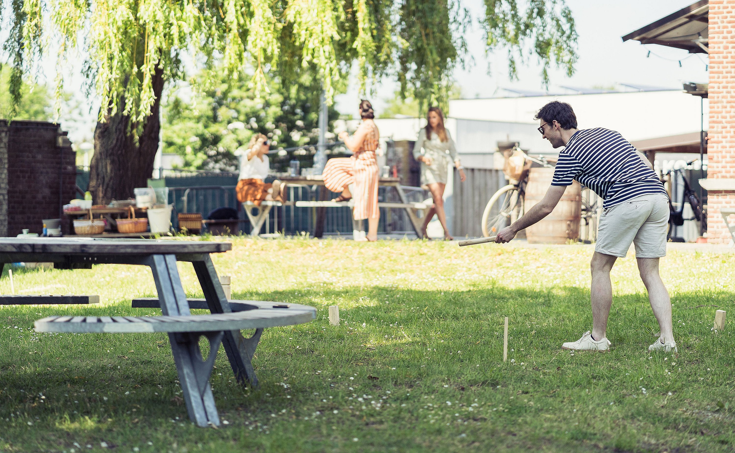 Man throwing a wooden baton during a lawn game while other guests stand and watch near the picnic setup.