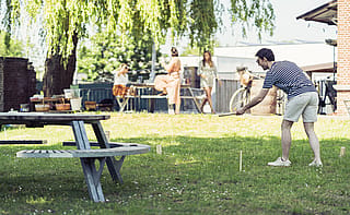 Man throwing a wooden baton during a lawn game while other guests stand and watch near the picnic setup.