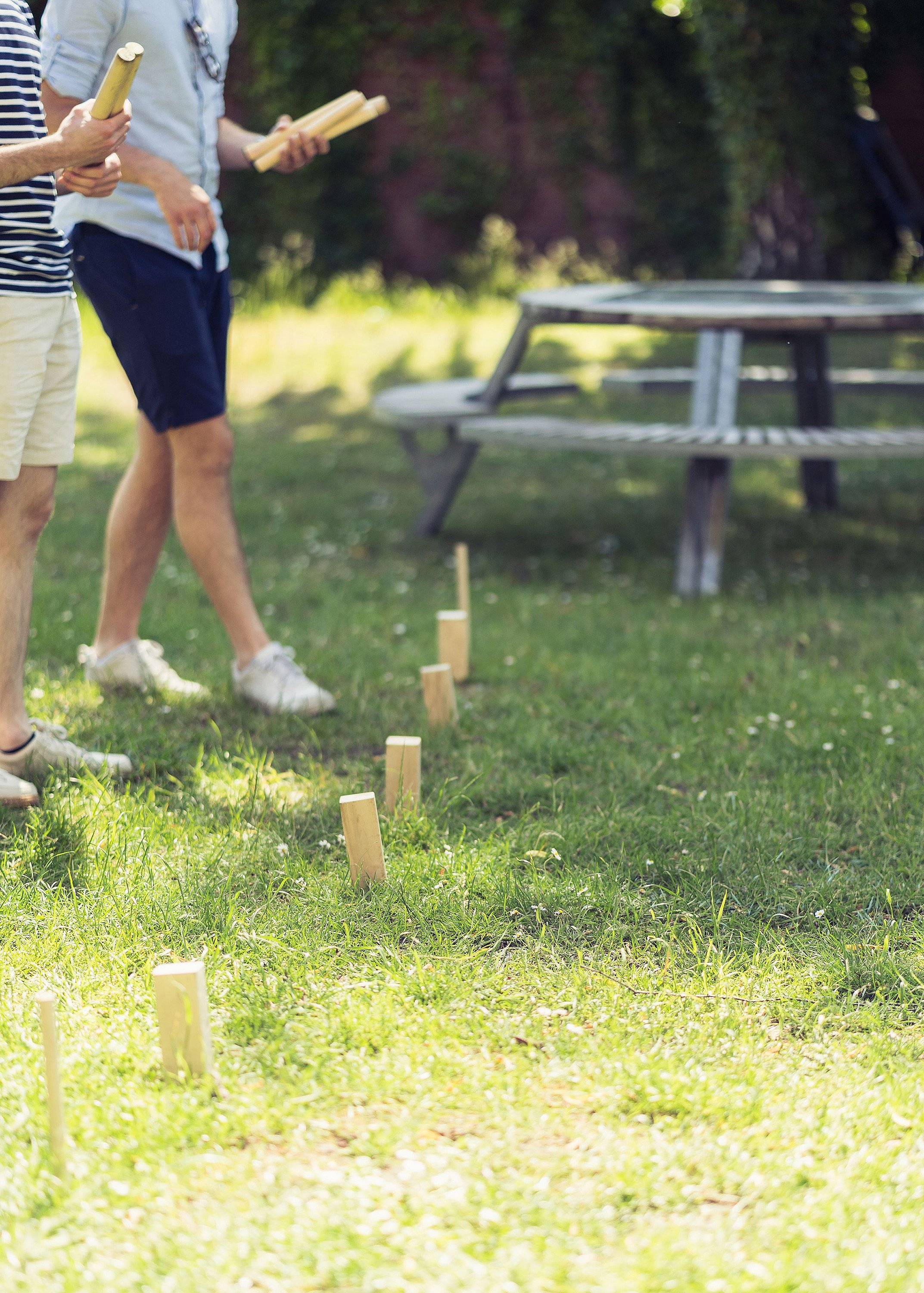 Close-up of a Kubb game set on the grass with players holding wooden batons and a picnic table blurred in the background.