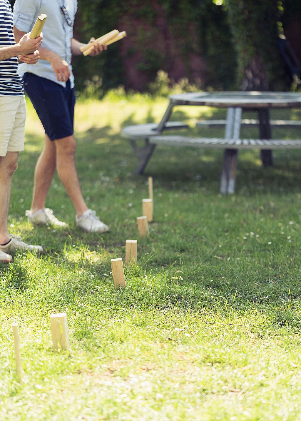 Close-up of a Kubb game set on the grass with players holding wooden batons and a picnic table blurred in the background.