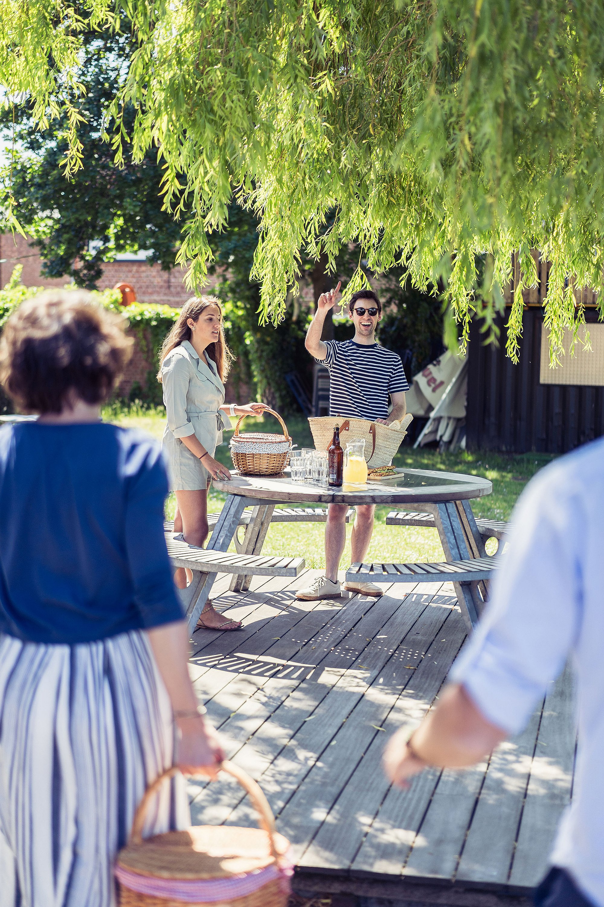 Two people standing by a round picnic table under a willow tree while friends approach carrying picnic baskets across the lawn.