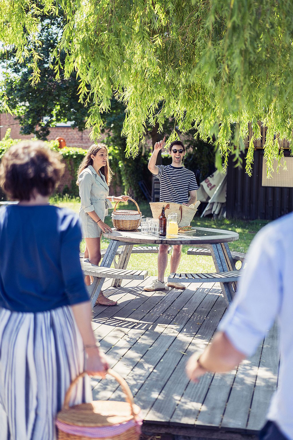 Two people standing by a round picnic table under a willow tree while friends approach carrying picnic baskets across the lawn.