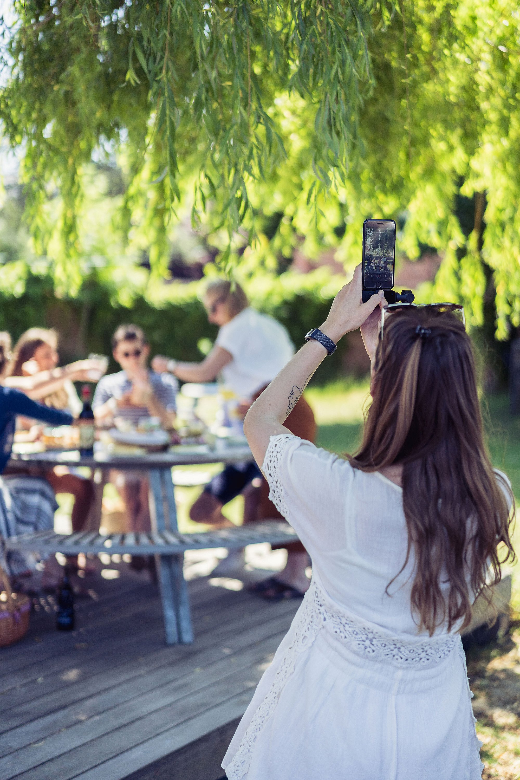 Woman photographing friends at the picnic with her phone while they sit together under hanging branches.