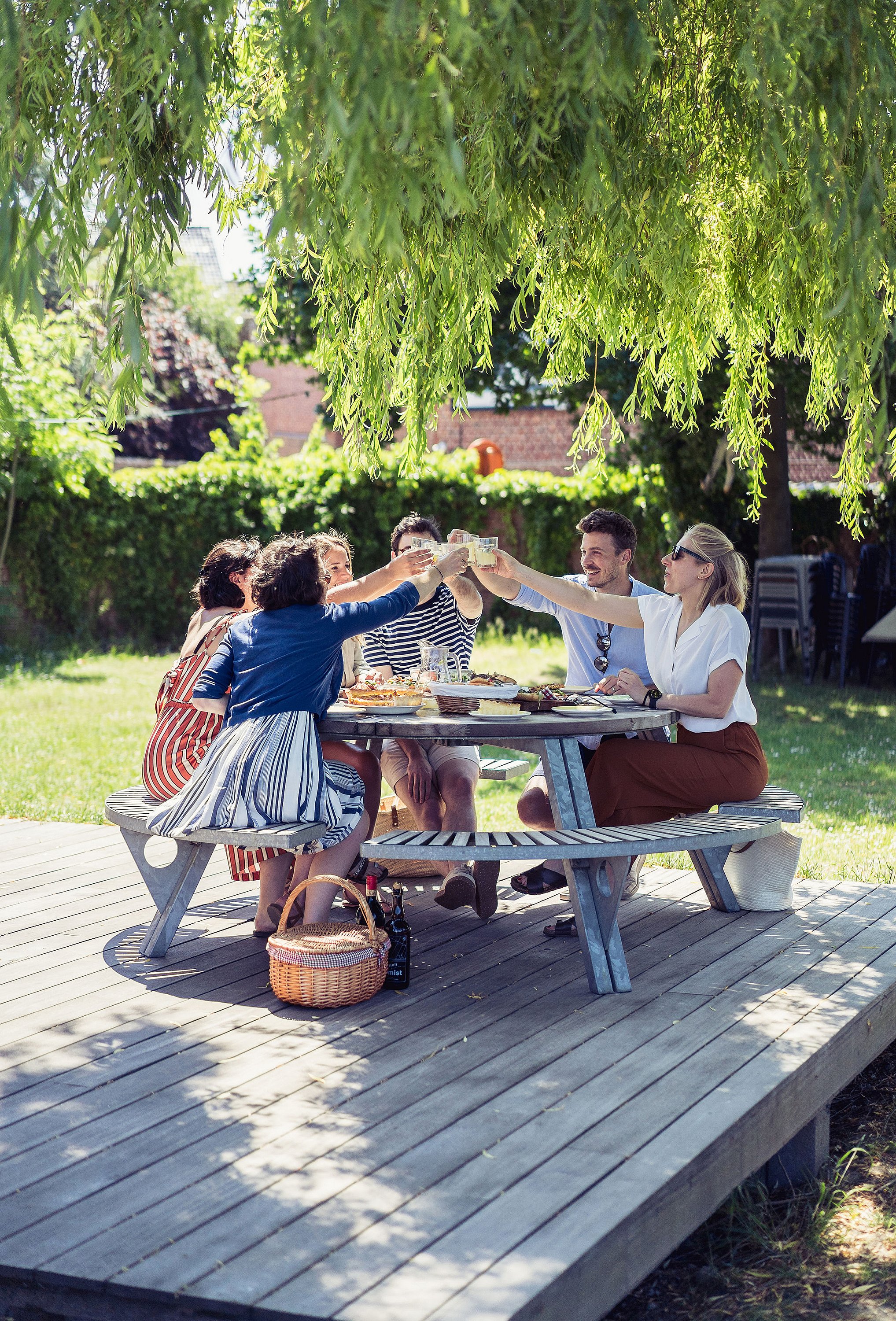 Group of friends toasting with drinks around a round picnic table under a leafy willow tree.