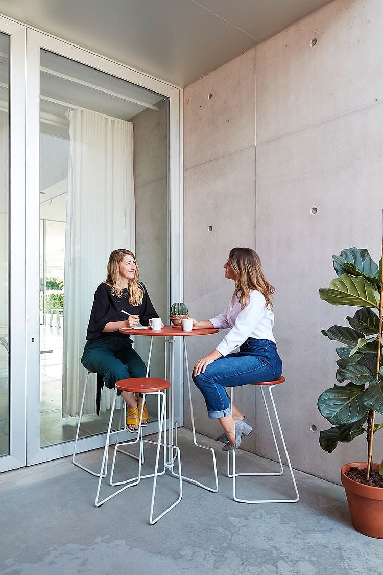 Two women having a casual meeting at a tall round outdoor table with matching stools on a sheltered concrete patio beside a large potted plant.