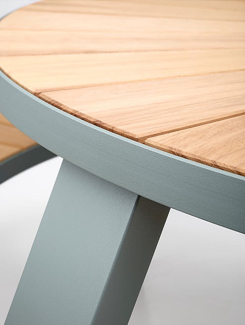   Détail d'un plateau de table rond en bois avec un rebord métallique vert pâle et un pied incliné, mettant en valeur le bord incurvé et l'assemblage.