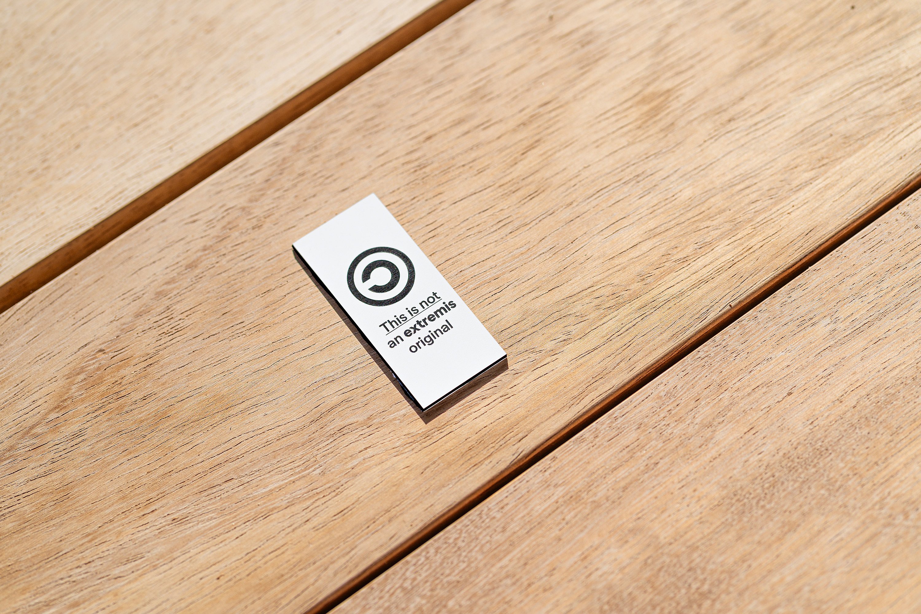 Petite étiquette métallique portant l'inscription « Ceci n'est pas une pièce originale Extremis » apposée sur un plateau de table en bois à lattes.