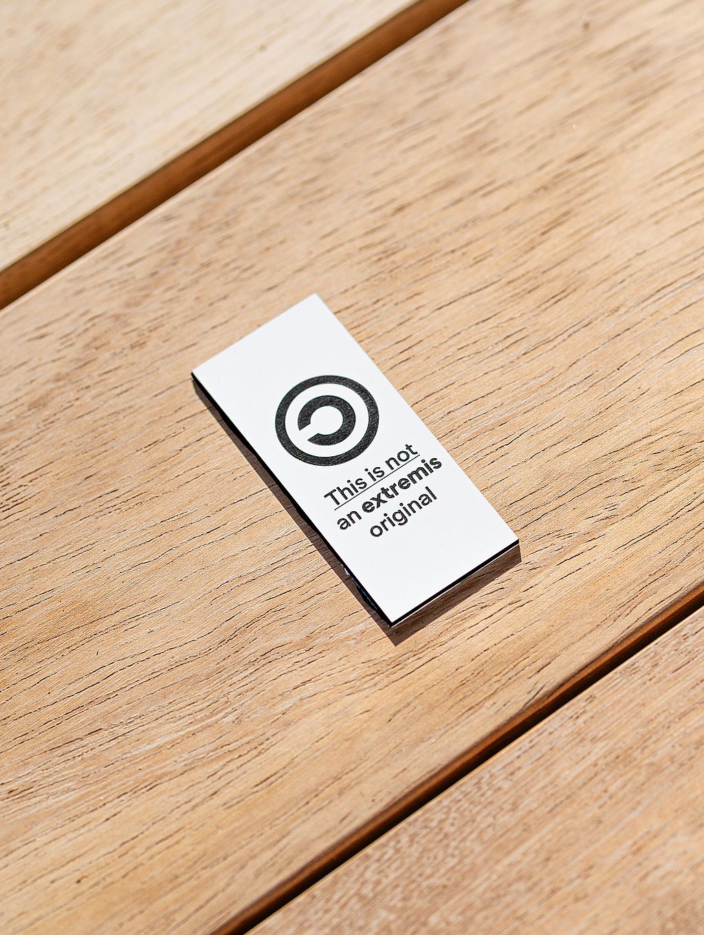 Small metal tag reading “This is not an extremis original” placed on a slatted wooden tabletop