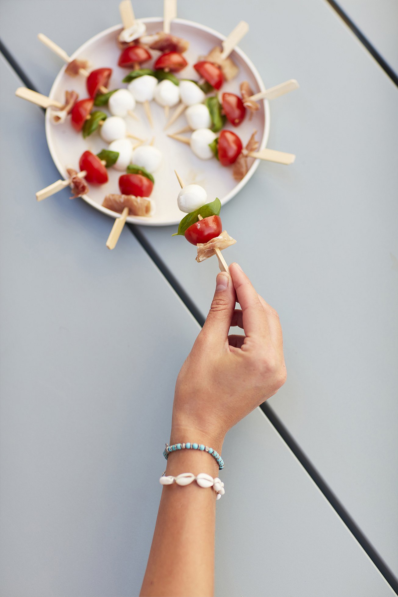 Main tenant une brochette de mozzarella, tomate, basilic et jambon sec au-dessus d’une table bleu pâle, à côté d’une assiette de brochettes assorties.