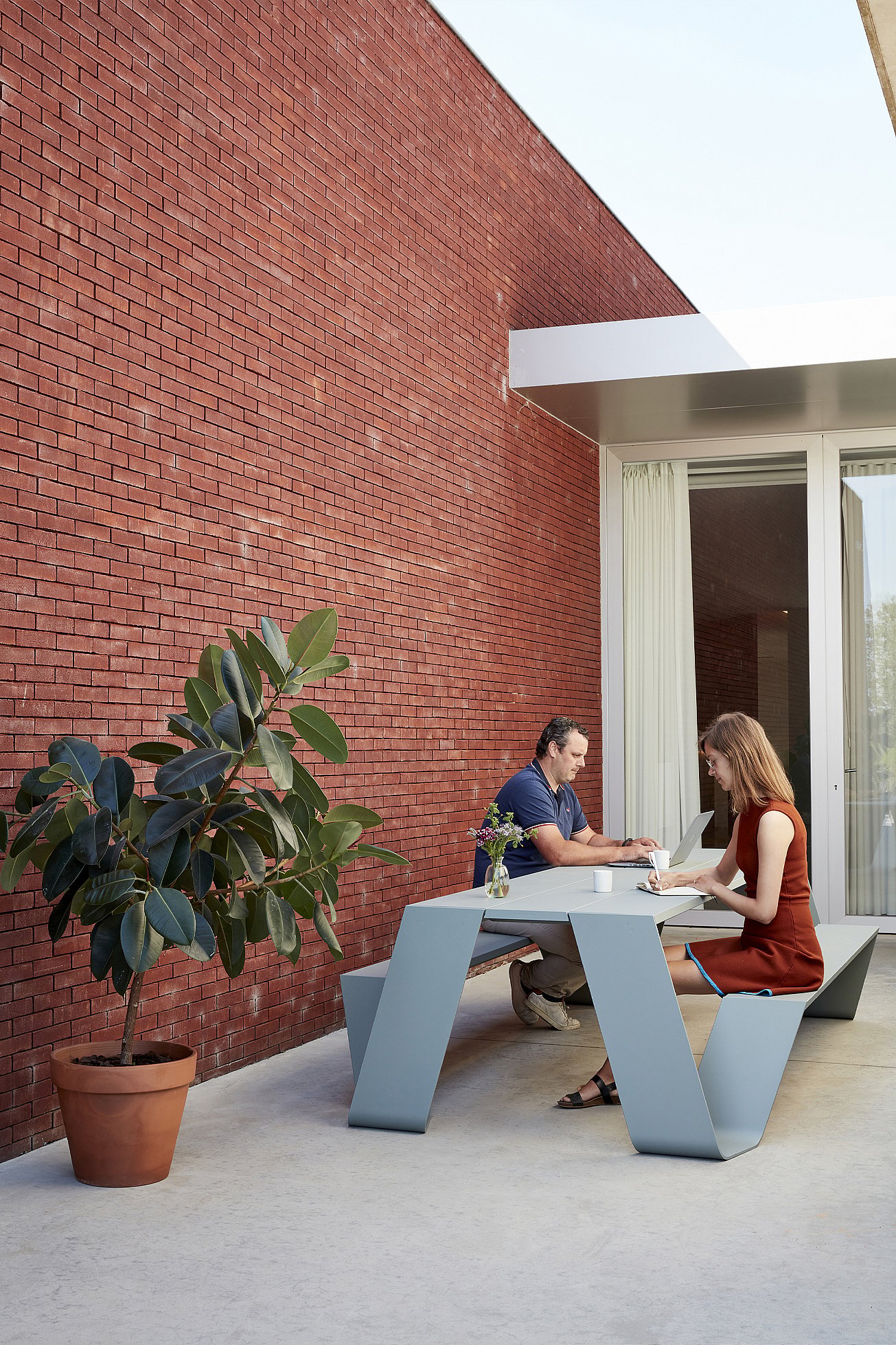 Deux personnes sont assises l’une en face de l’autre à une table Hopper AA bleu pâle sur une terrasse à côté d’un mur de briques rouges, avec un caoutchouc en pot au premier plan.