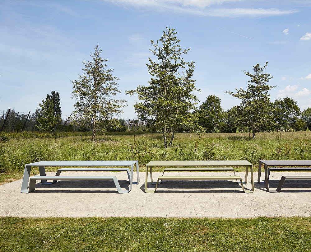 Trois tables de pique-nique tout en aluminium Hopper AA dans différentes couleurs sourdes se tiennent côte à côte sur un chemin de gravier dans un paysage herbeux ouvert avec des arbres sous un ciel bleu.
