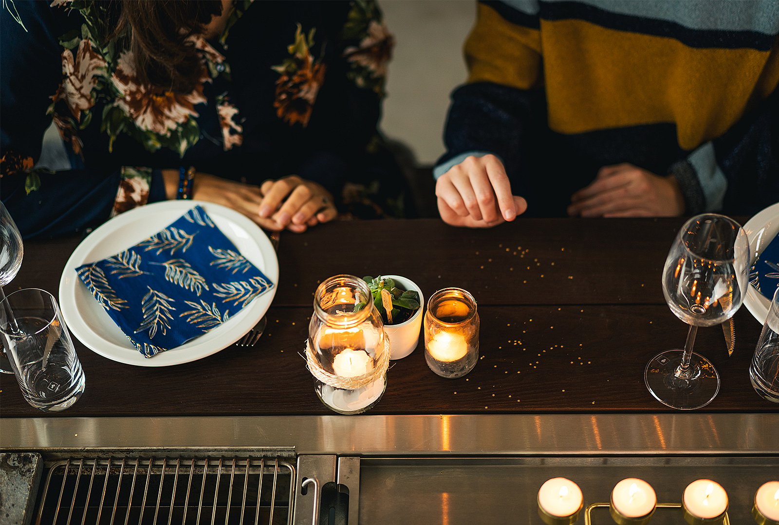 Warme Draufsicht auf einen dunklen Hopper-Holztisch für ein gemütliches Essen mit Kerzen, einer gefalteten blauen Serviette und zwei Personen, die neben dem eingebauten Grill sitzen.