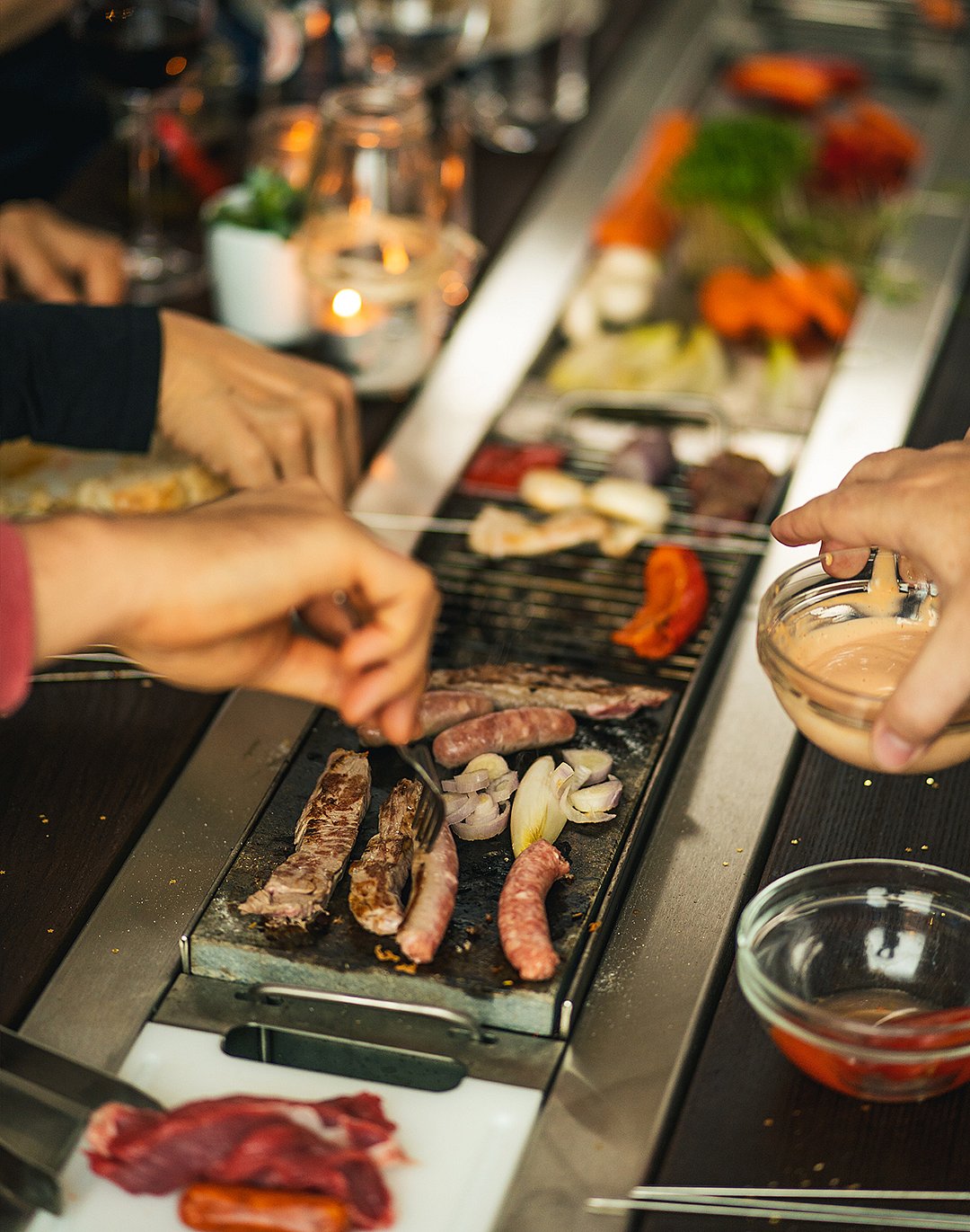 Nahaufnahme von Würstchen, Fleisch, Zwiebeln und Gemüse, die auf dem Tischgrill brutzeln, während mehrere Hände darum herum kochen und Essen servieren.