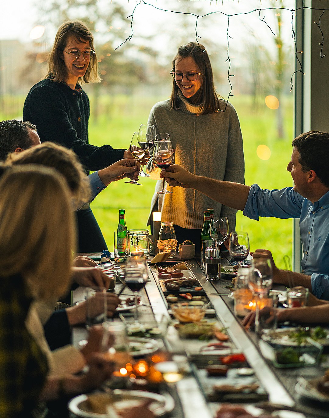 Freunde rund um den Hopper-Tisch heben Weingläser für einen Toast während eines festlichen Abendessens im Innenbereich an einem großen Fenster mit Blick auf eine grüne Landschaft.