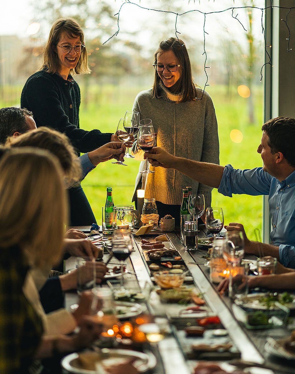 Freunde rund um den Hopper-Tisch heben Weingläser für einen Toast während eines festlichen Abendessens im Innenbereich an einem großen Fenster mit Blick auf eine grüne Landschaft.