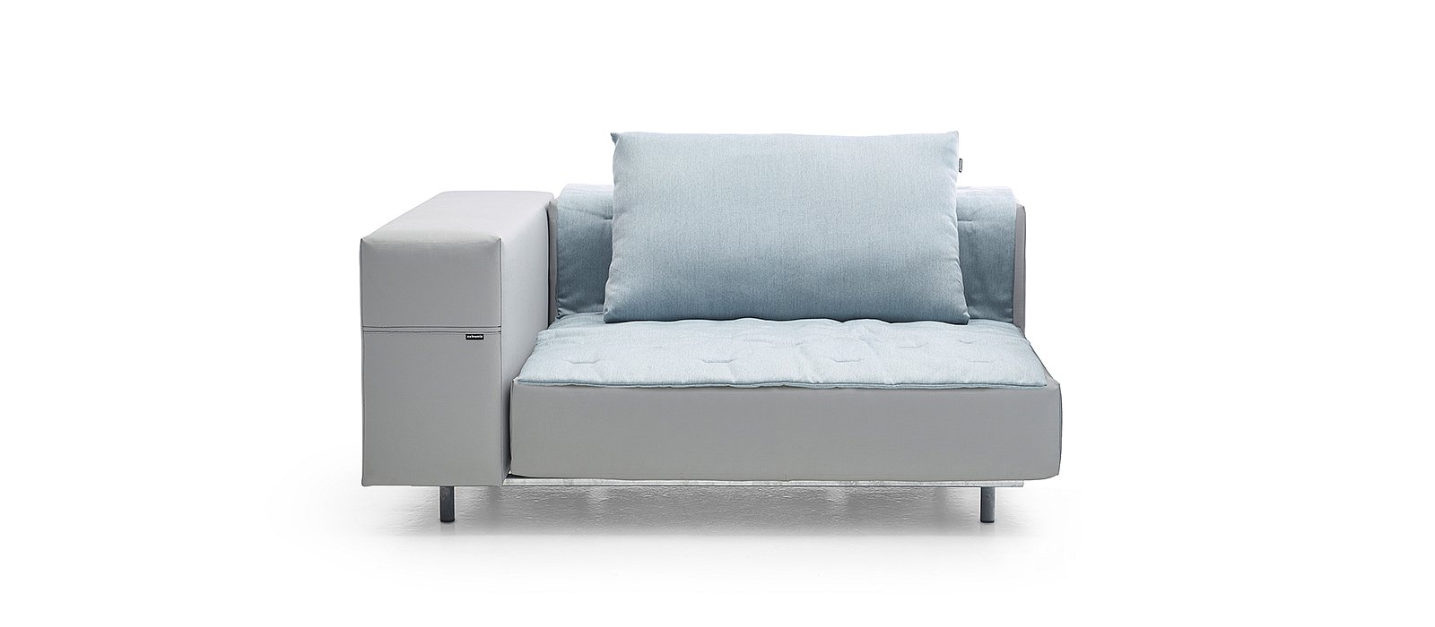 Salon extérieur aménagé au bord d’une plage rocheuse, avec un grand canapé modulable posé sur l’herbe pendant qu’une femme le vaporise avec un tuyau, un autre y verse un seau d’eau et un homme assis se détend sur les coussins.