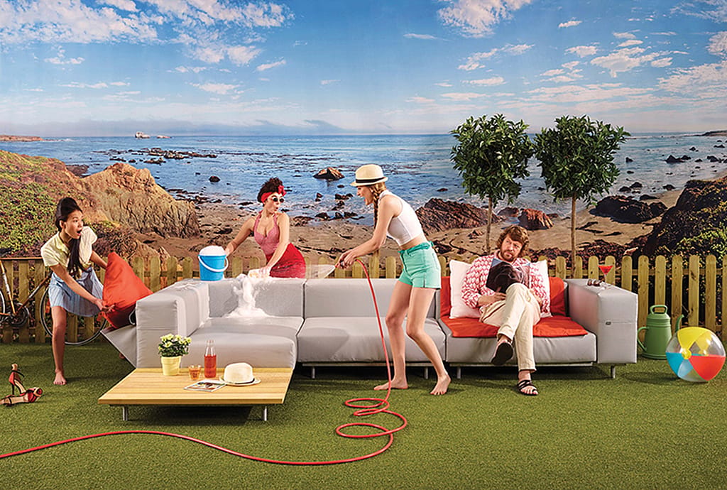 Salon extérieur aménagé au bord d’une plage rocheuse, avec un grand canapé modulable posé sur l’herbe pendant qu’une femme le vaporise avec un tuyau, un autre y verse un seau d’eau et un homme assis se détend sur les coussins.