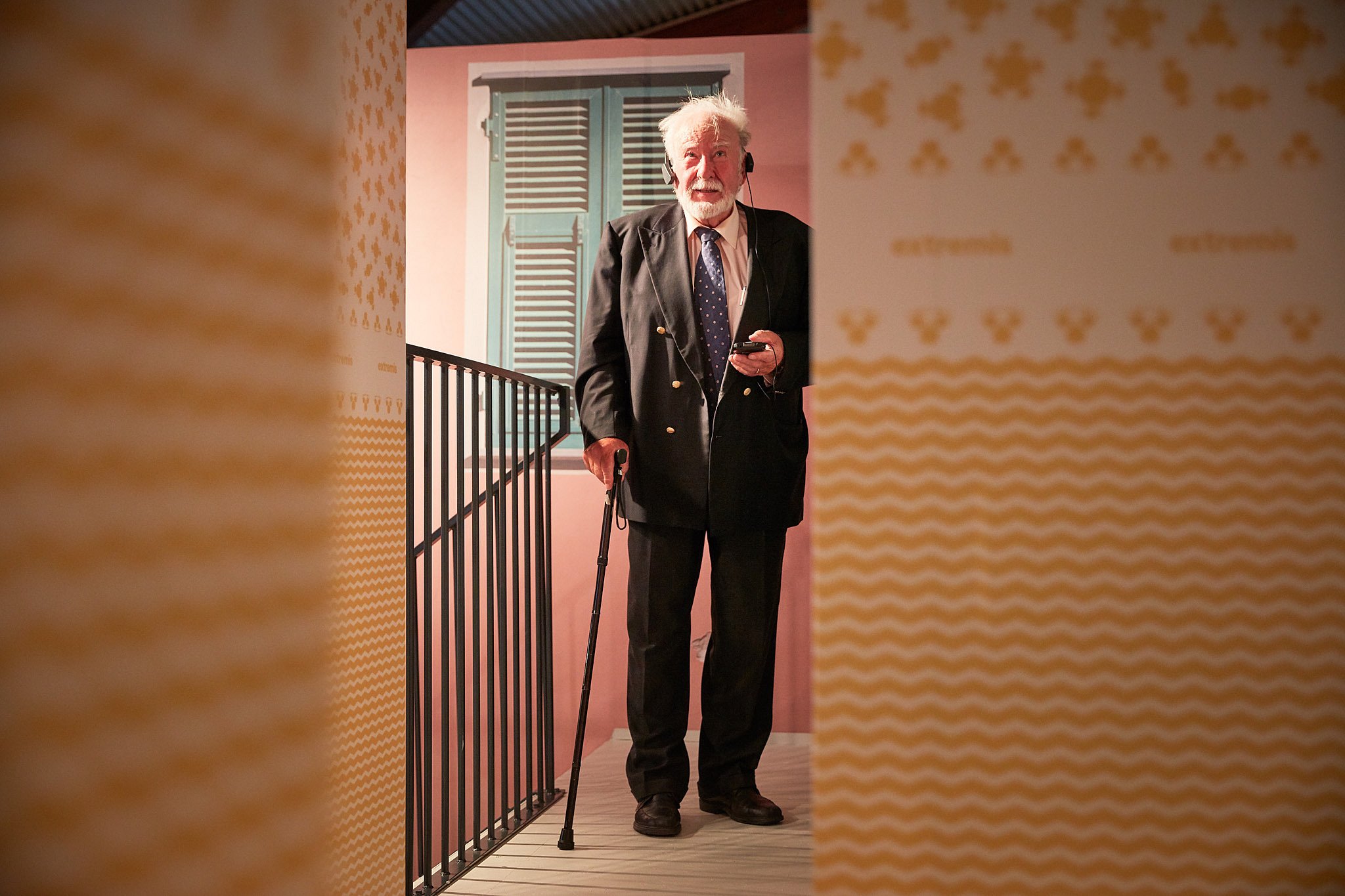Older man in a dark suit uses headphones and an audio guide while standing on the balcony walkway of the exhibition set.