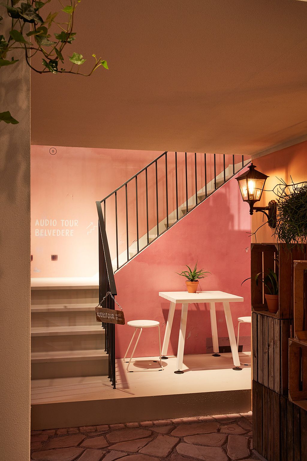 Quiet corner of the stand with pink walls, a white square table and stools under a staircase, lit like a small Mediterranean interior.