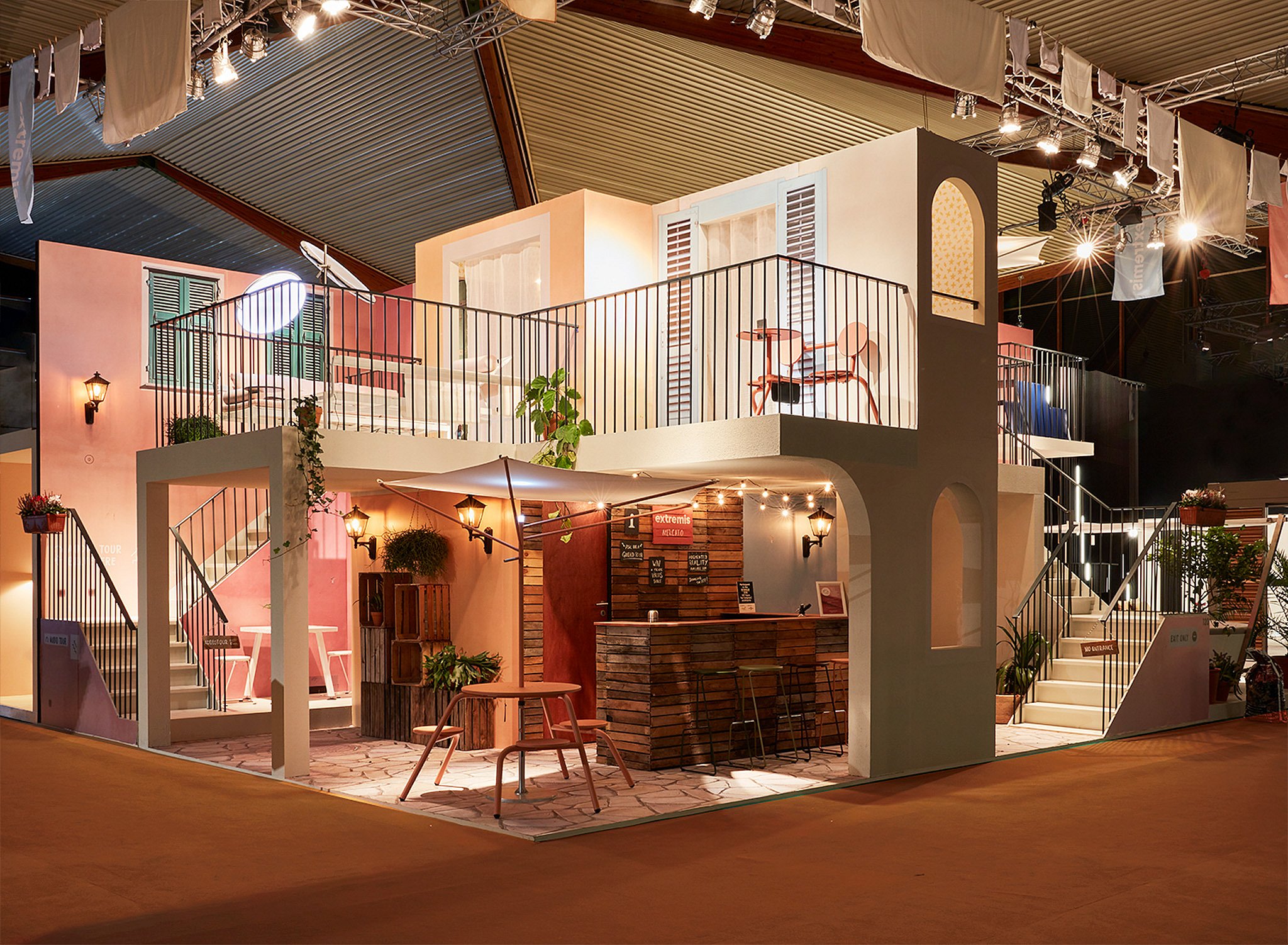 Full exhibition stand styled like a pastel Italian street, with balconies, shutters, stairs, string lights, and compact outdoor furniture arranged across two levels inside the fair hall.