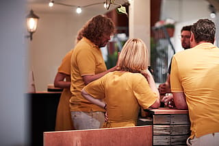 Group of people in coordinated yellow clothing gather and talk around a rustic wooden bar beneath string lights.