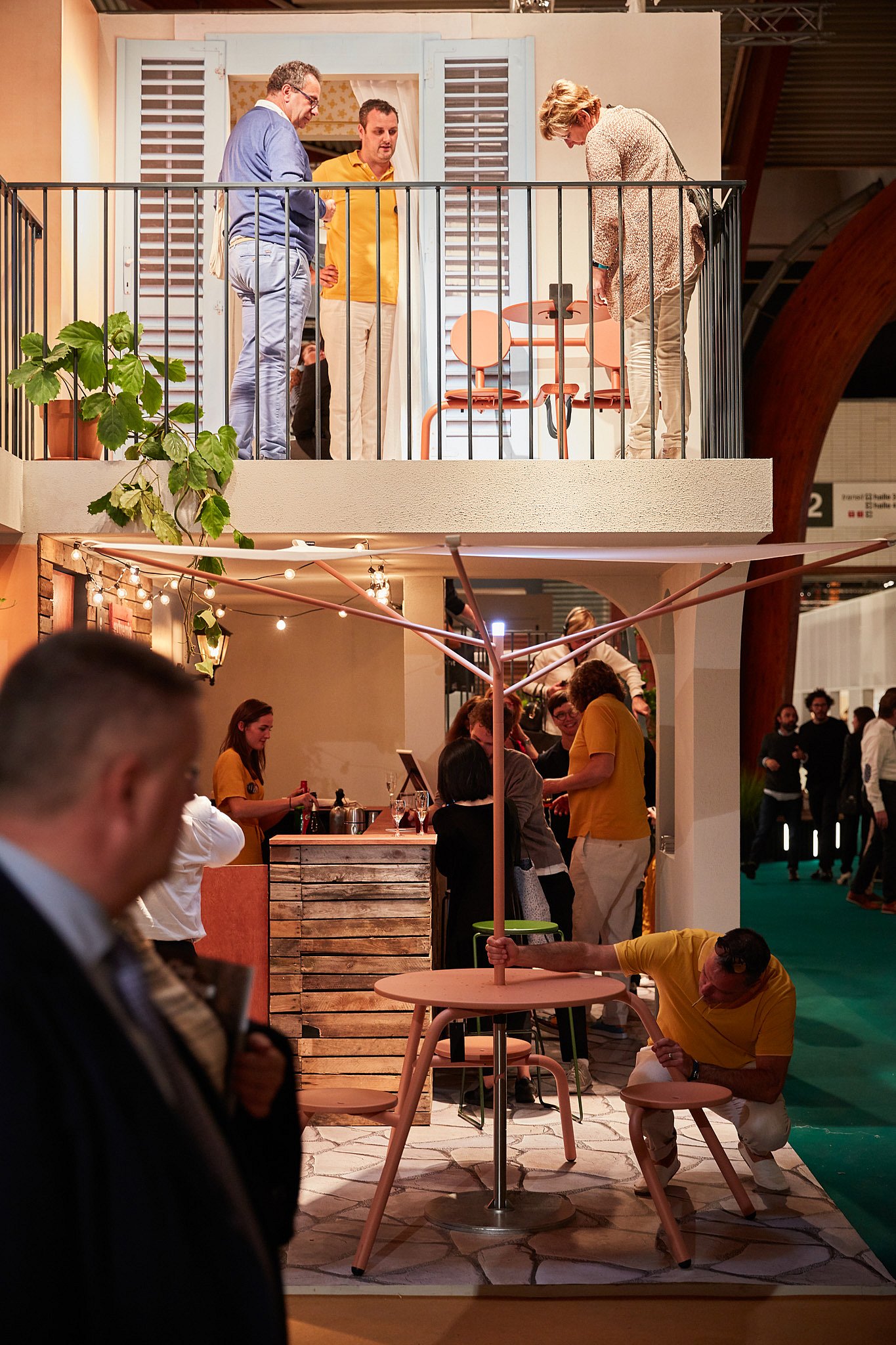 Busy view of the stand with visitors exploring the balcony and ground floor while a pink Acacia parasol and Bistroo set fill the foreground.