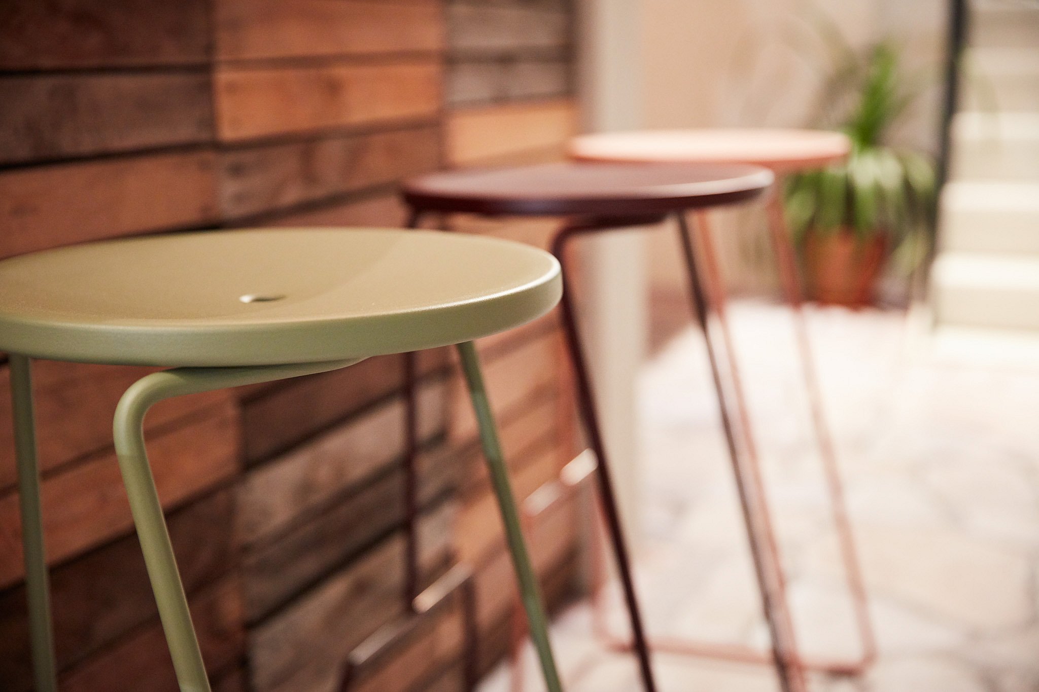 Three slim round-topped stools in green, dark brown, and pale pink stand in a row beside a wooden wall.