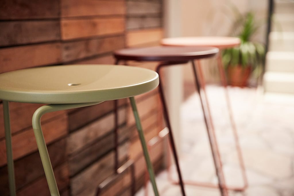 Three slim round-topped stools in green, dark brown, and pale pink stand in a row beside a wooden wall.