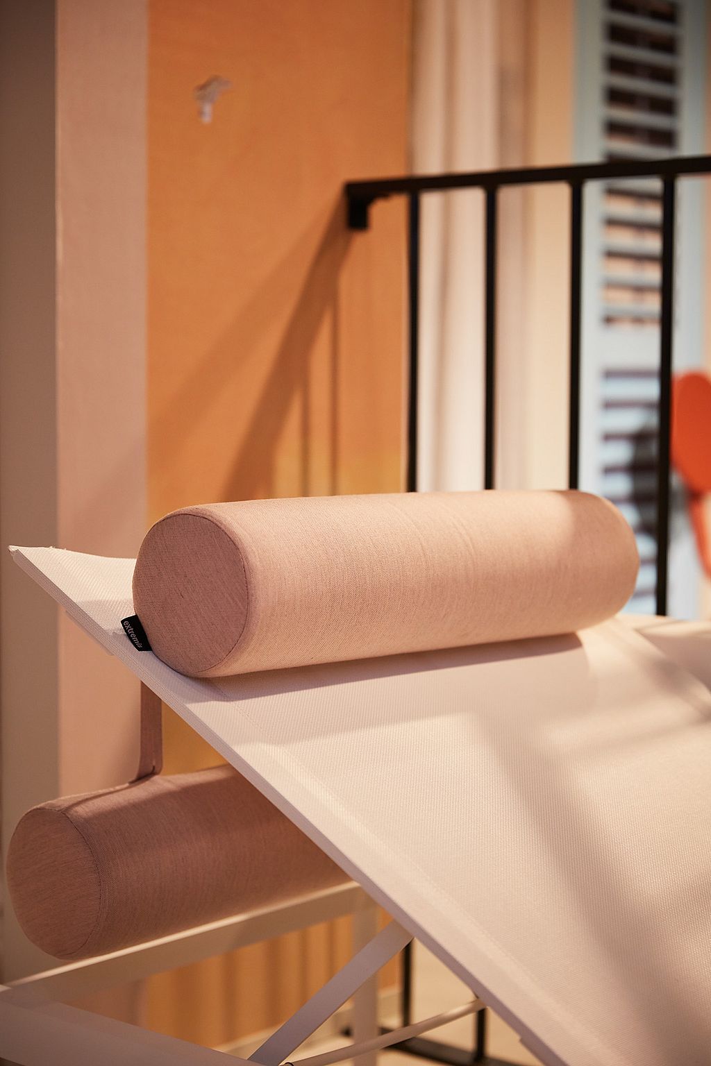 Close-up of a pale cylindrical cushion resting on an angled white outdoor lounger frame with black railing in the background.