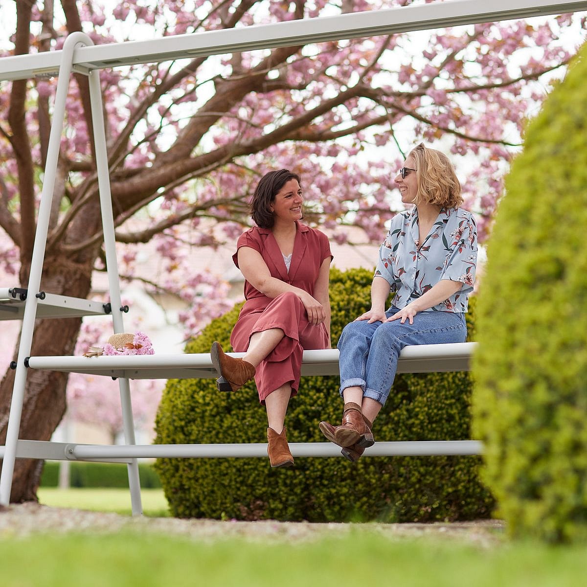 Deux femmes assises sur une structure blanche AMAi (table et banc) sous des arbres en fleurs roses se sourient dans un jardin paysager.