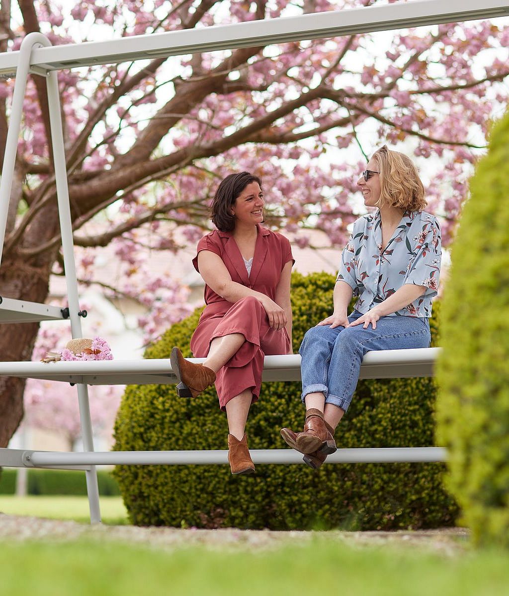 Deux femmes assises sur une structure blanche AMAi (table et banc) sous des arbres en fleurs roses se sourient dans un jardin paysager.