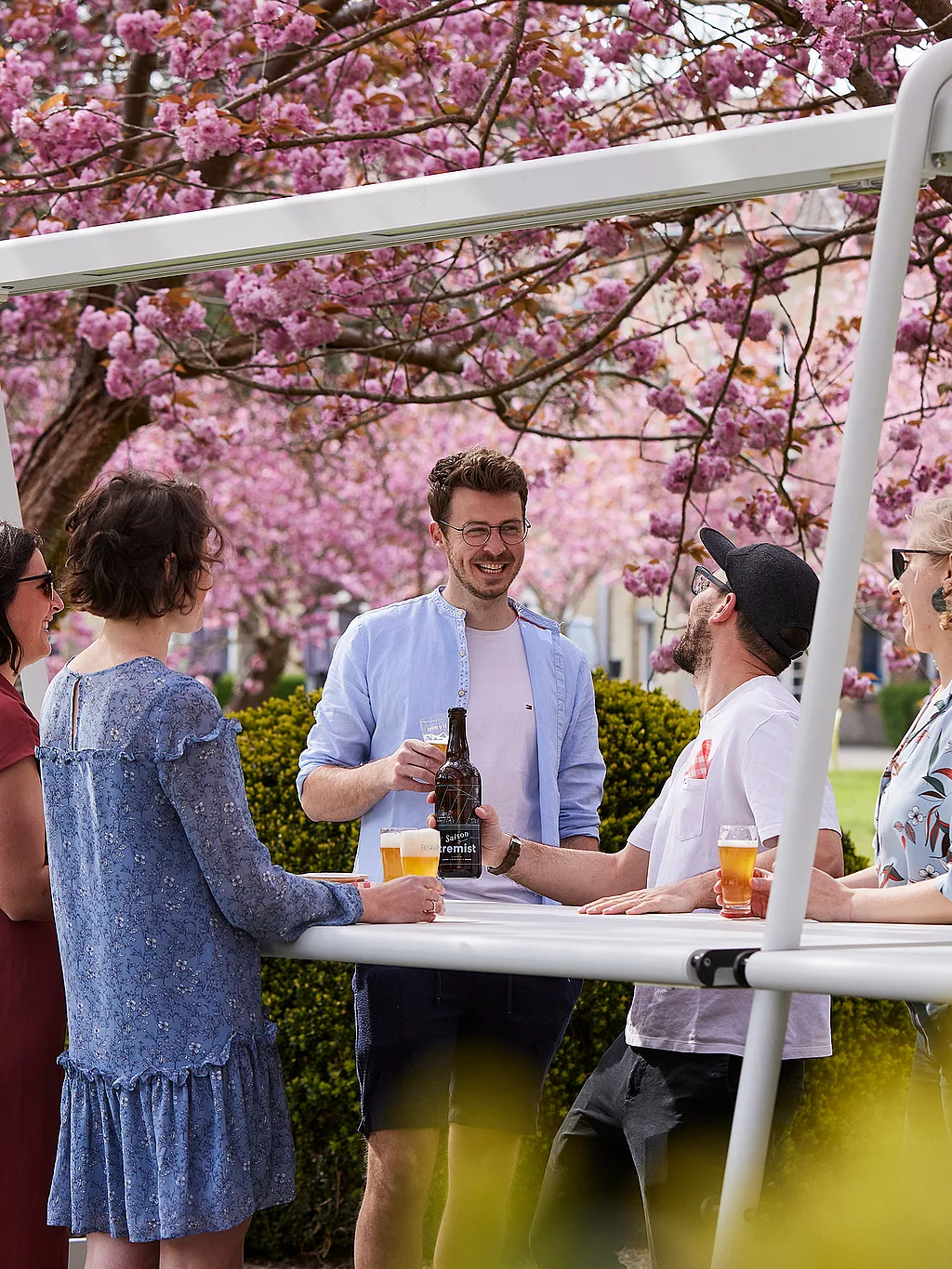 Cinq adultes réunis autour d'une table haute blanche sous des cerisiers en fleurs, bières à la main, discutent en plein air.