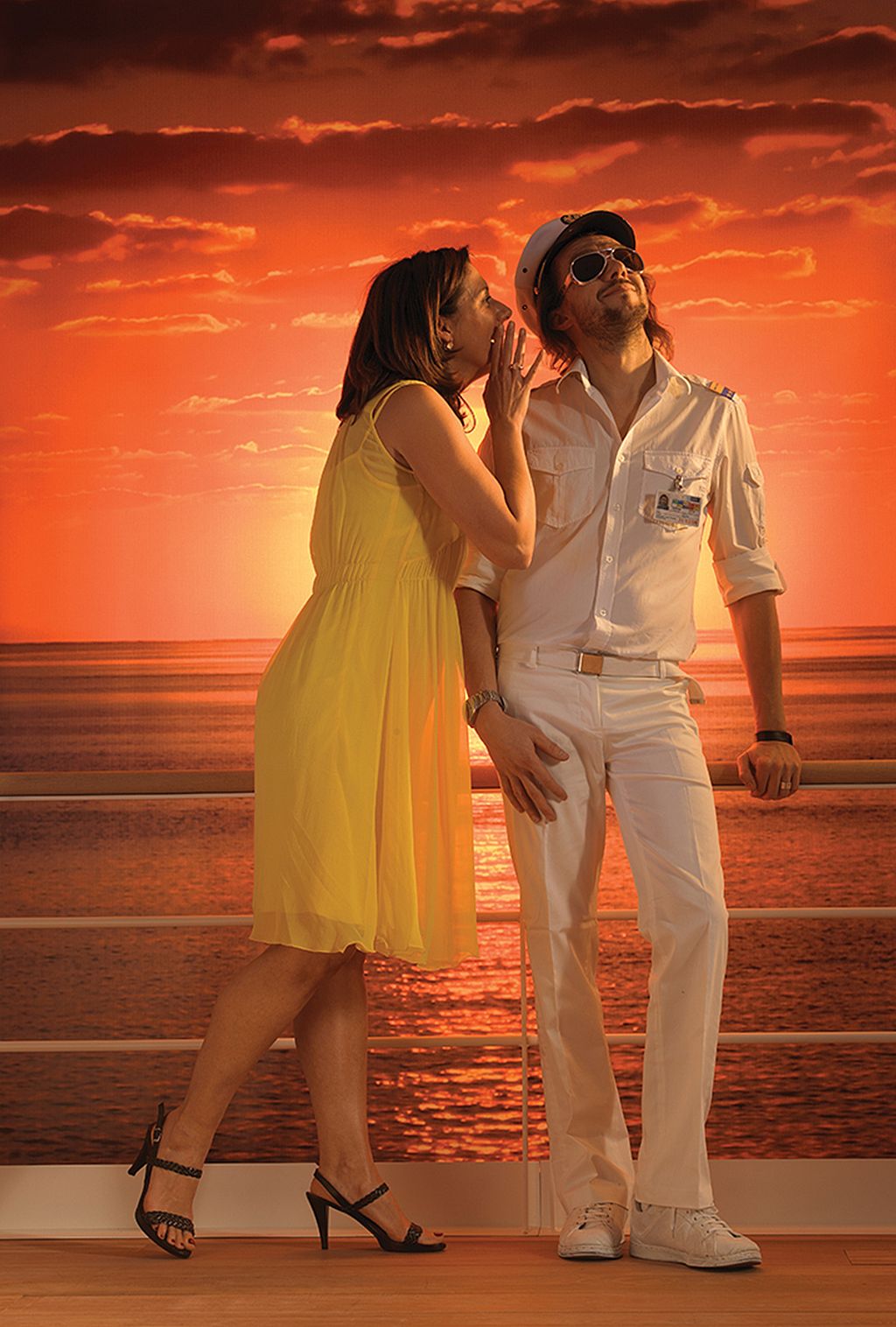 Eine Frau in einem gelben Kleid beugt sich zu einem Mann, der wie ein Yachtkapitän in weißer Uniform und Sonnenbrille gekleidet ist und hinter dem sich ein leuchtend orangefarbener Sonnenuntergang über dem Meer befindet.