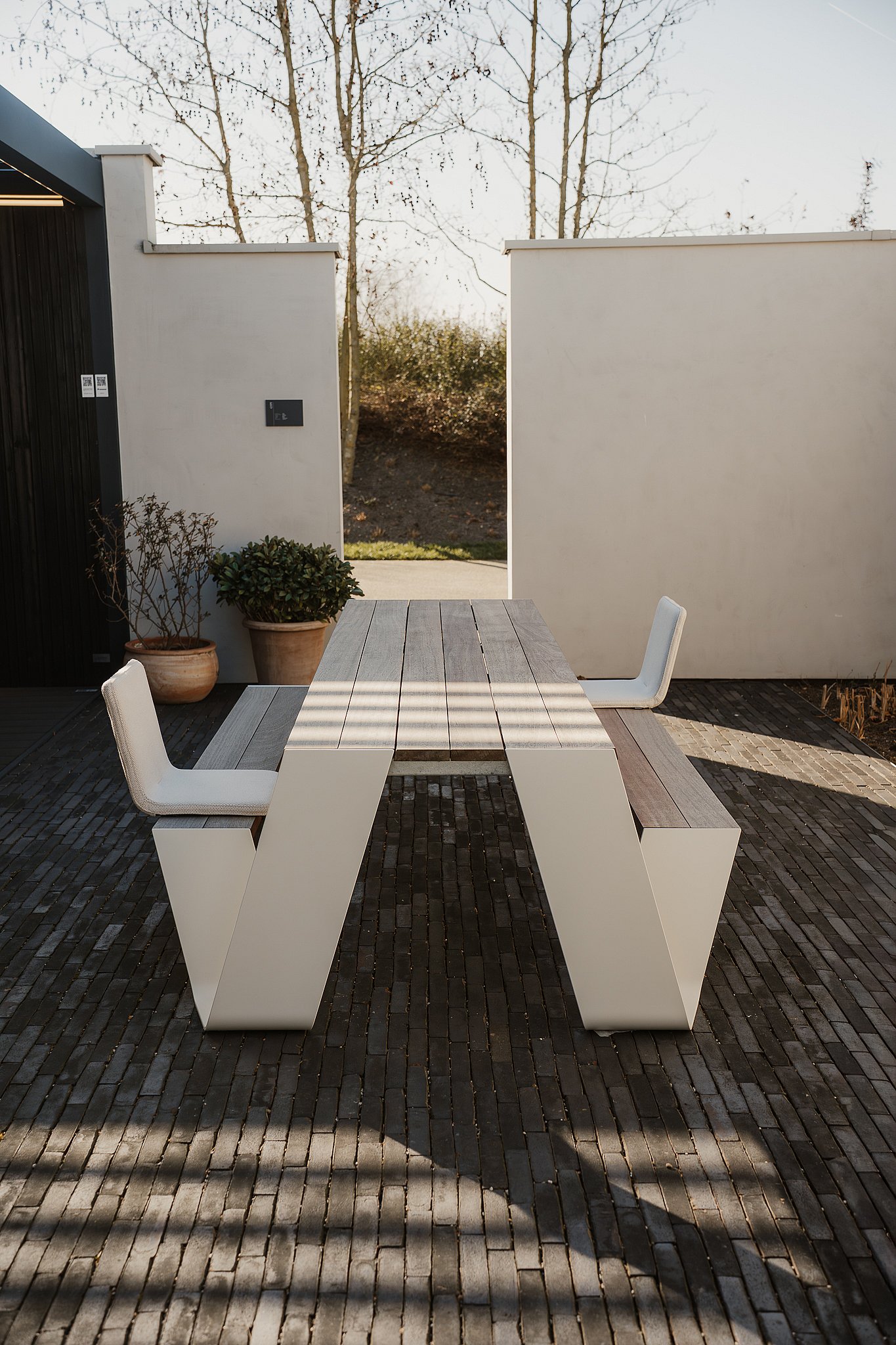   Vue latérale d'une table de pique-nique Hopper avec deux chaises BYOS fixées à ses extrémités, installée dans une cour en briques entre des murs blancs et des plantes en pot.