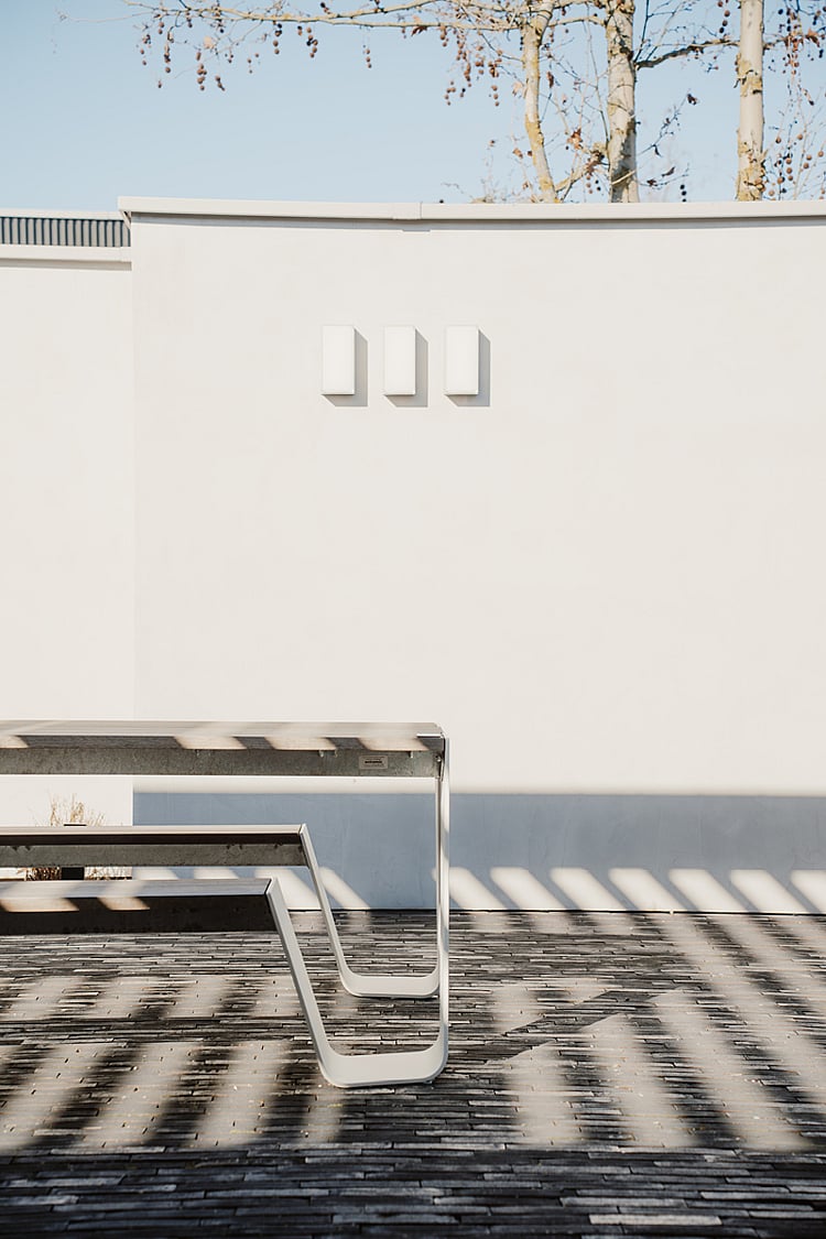   Vue latérale d'une table et d'un banc Hopper aux pieds en métal courbés, adossés à un mur blanc uni. L'ombre d'une pergola s'étend sur le pavage.