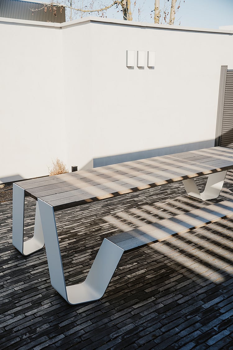 Table de pique-nique Hopper vide, aux lattes de bois patinées et aux pieds en métal blanc, sur une terrasse en briques. Des ombres rayées se projettent sur la table et le sol.