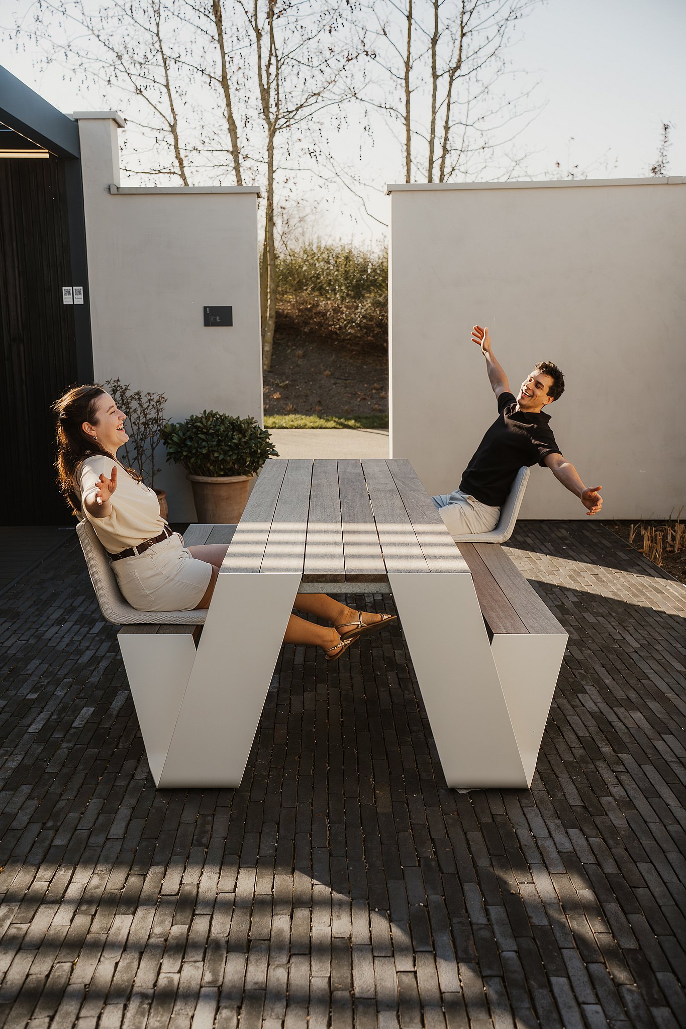Deux personnes se prélassent en arrière sur des sièges BYOS rembourrés, fixés de part et d'autre d'une table de pique-nique Hopper blanche, sur une terrasse en briques ensoleillée.