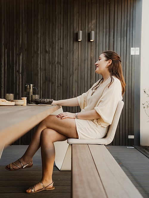   Une femme est confortablement installée sur une chaise BYOS à une table Hopper, sur une terrasse en bois sombre. De la vaisselle et des verres sont posés sur la table.