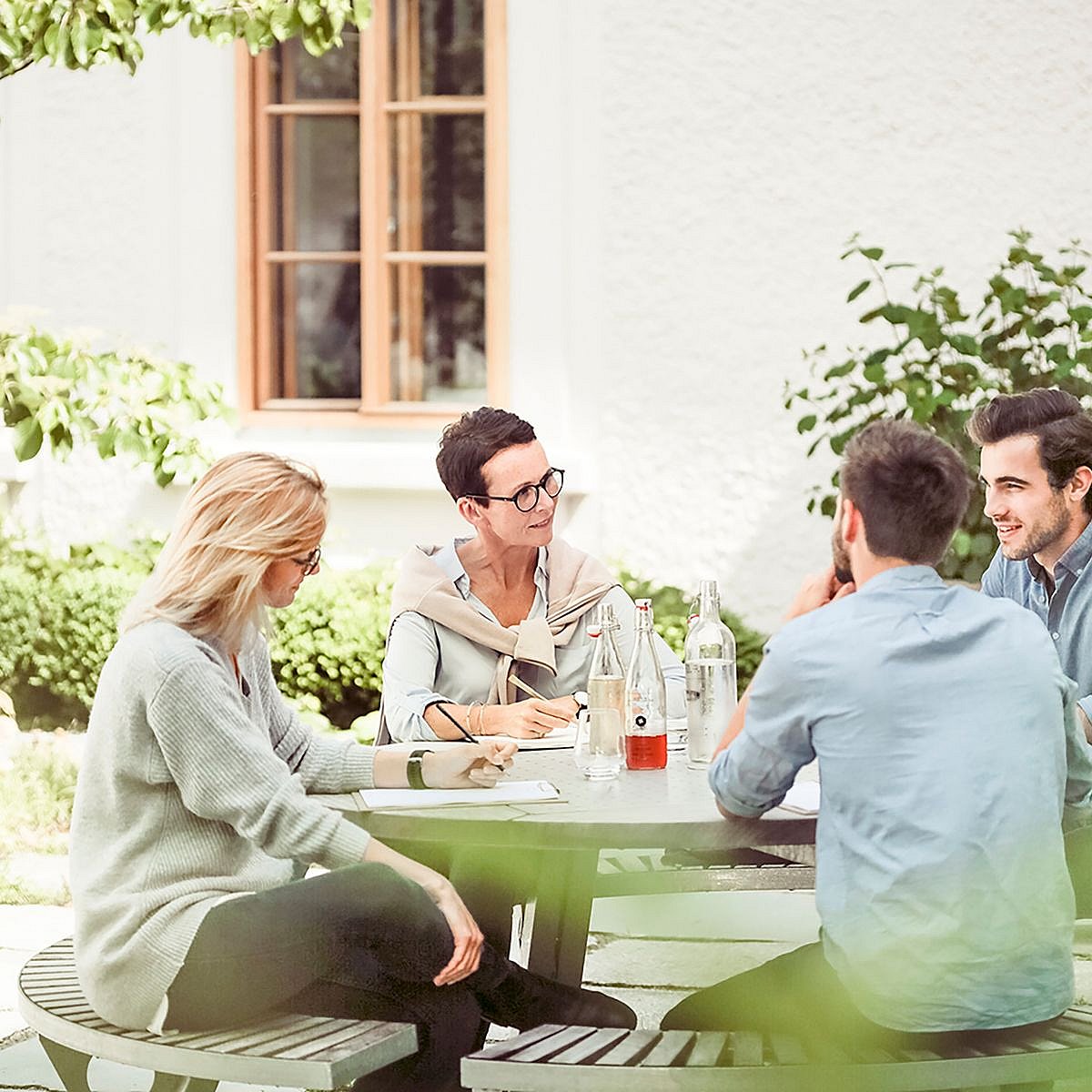 Four coworkers seated around a round outdoor table in a leafy courtyard, talking and taking notes beside glass bottles and notebooks.