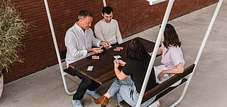 Eenvoudige lijntekening van vier mensen die rond een tafel zitten, met één spraakbel met kaartpakken en een andere met boze symbolen om aan te geven dat spelers hun kaarten niet mogen bespreken.
